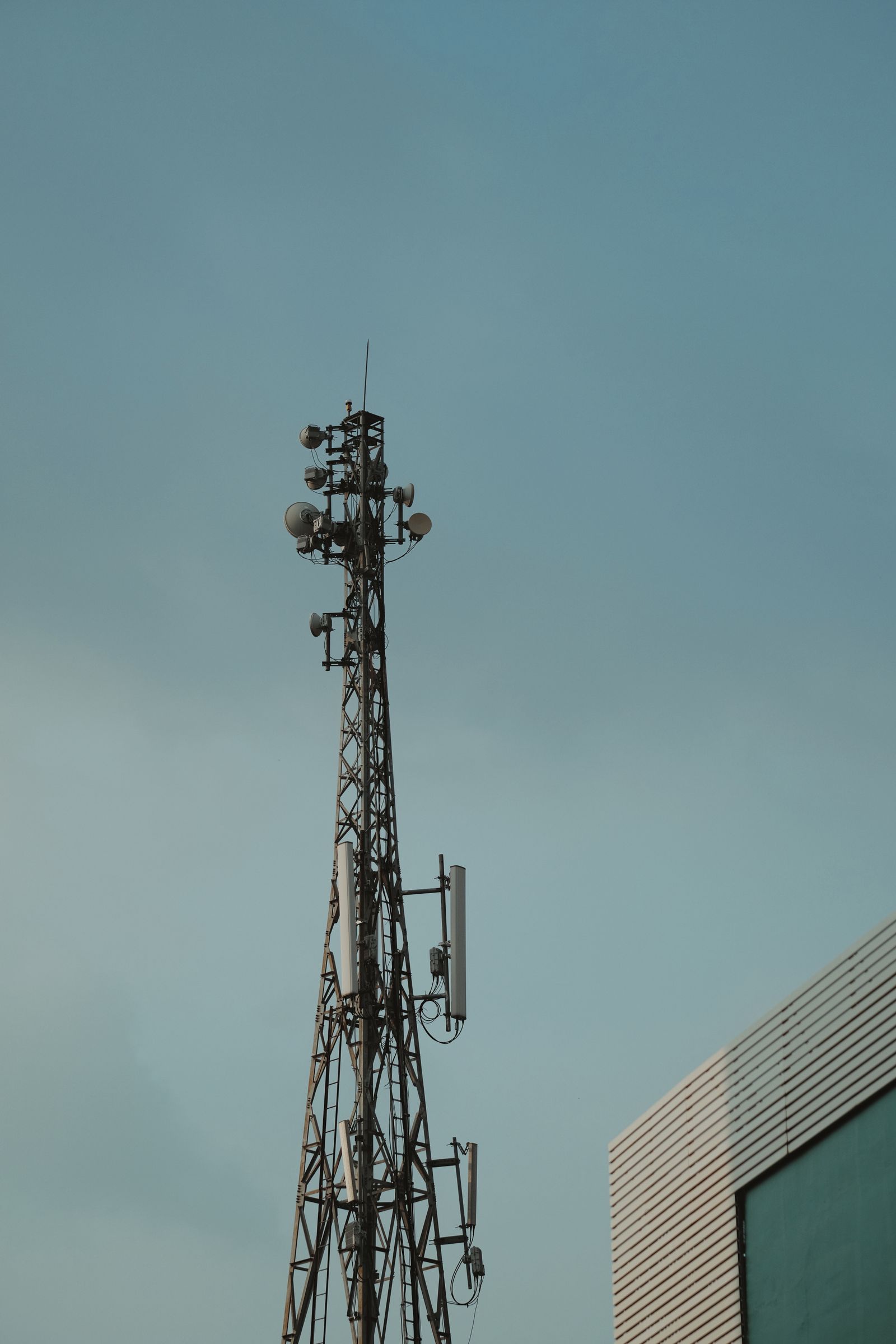 Torre de telecomunicações com antenas e parabólicas contra um céu azul-acinzentado, com a borda de um prédio à direita.