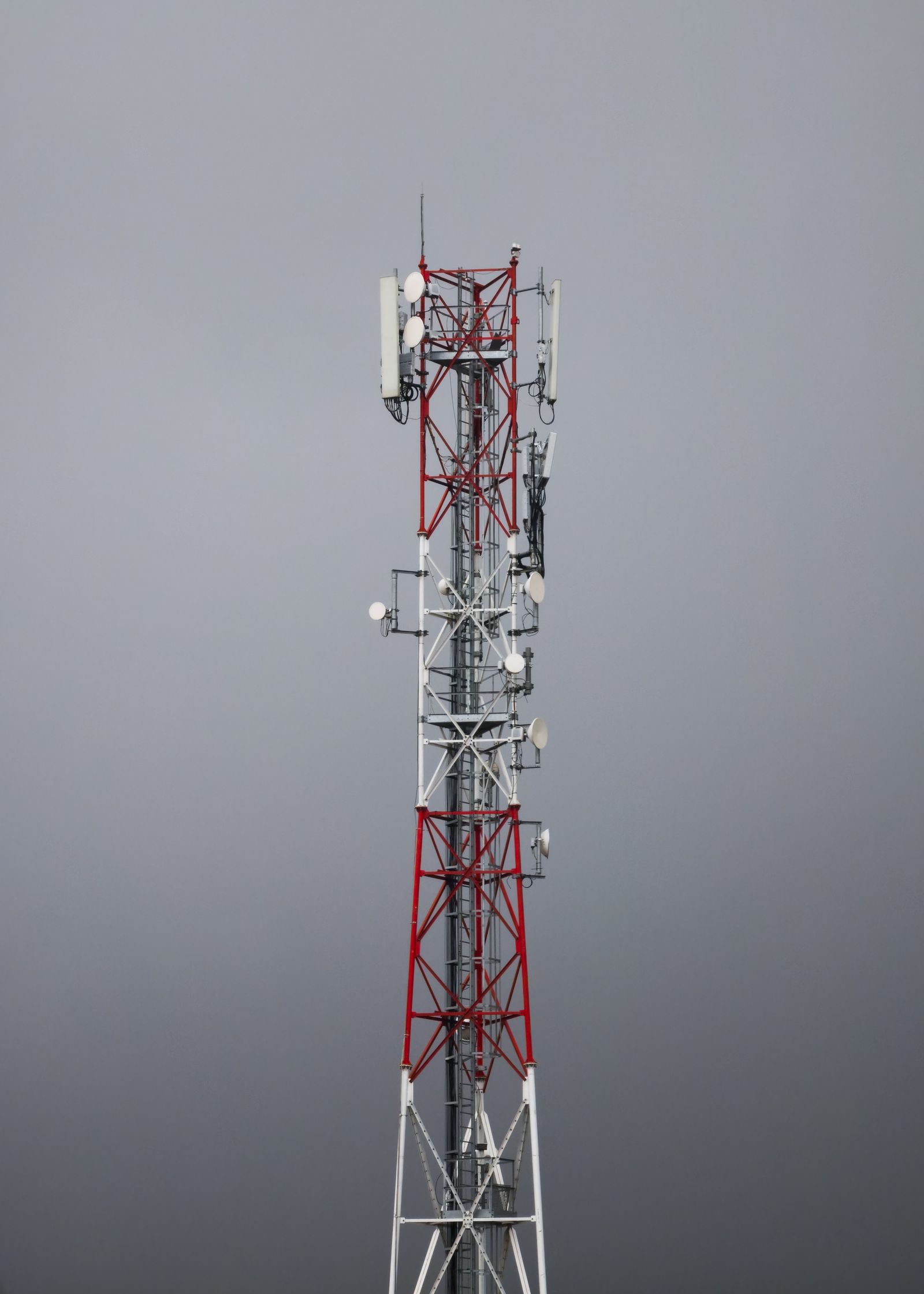 Torre de telecomunicações vermelha e branca com parabólicas contra um céu cinza.