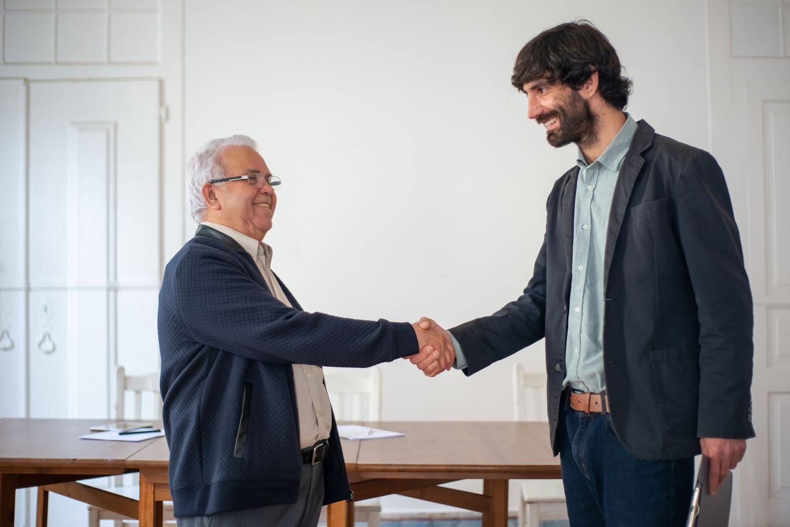 Dois homens apertando as mãos e sorrindo em uma sala clara com uma mesa de madeira.